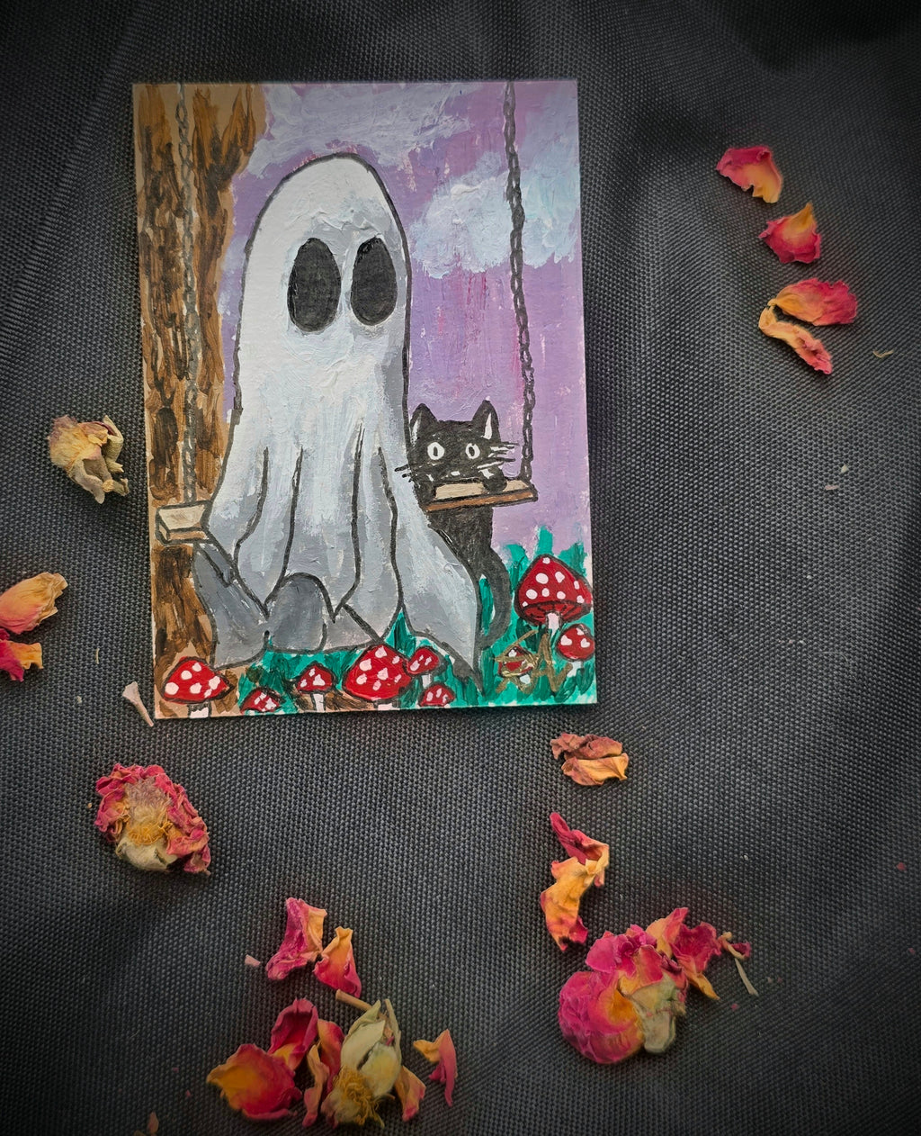 Original Ghost and Kitty ACEO painting featuring a white ghost, black cat on a swing, and red mushrooms, photographed on black fabric with dried rose petals.