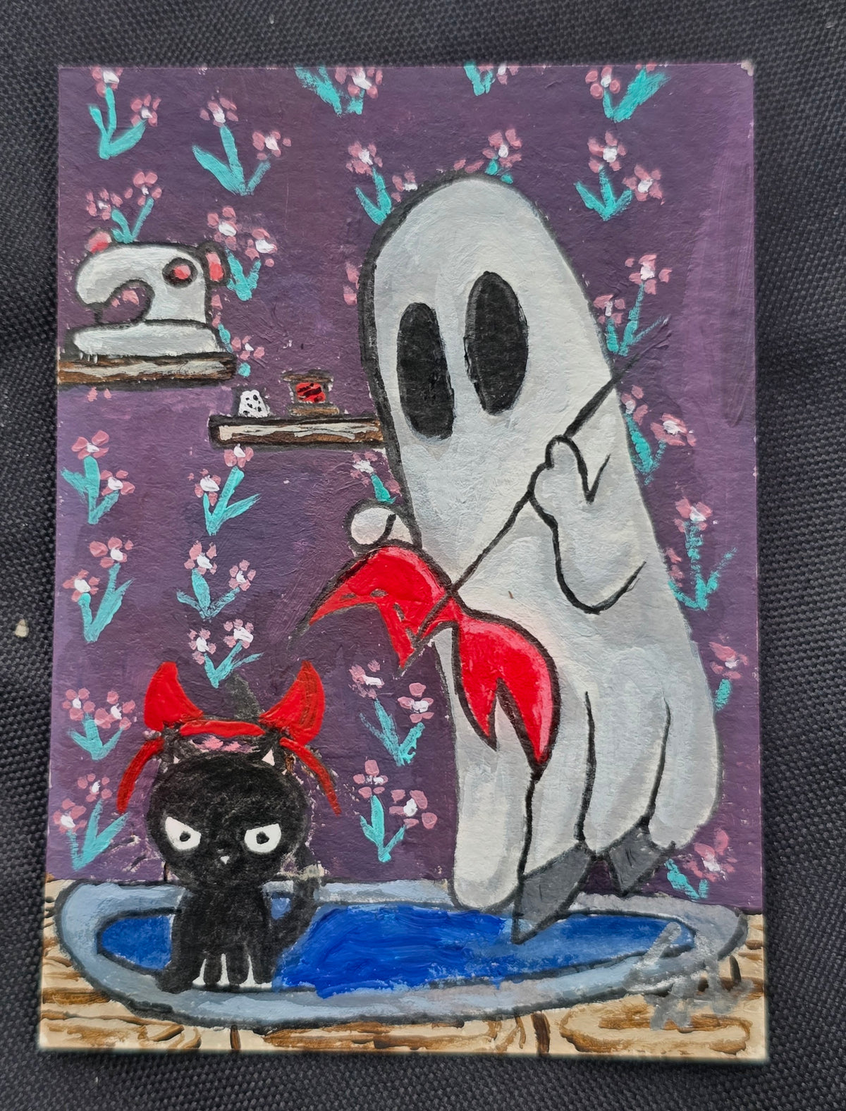 Original miniature painting of a white ghost sewing a red costume while a black cat wears red devil horns, set against purple floral wallpaper with sewing supplies nearby.