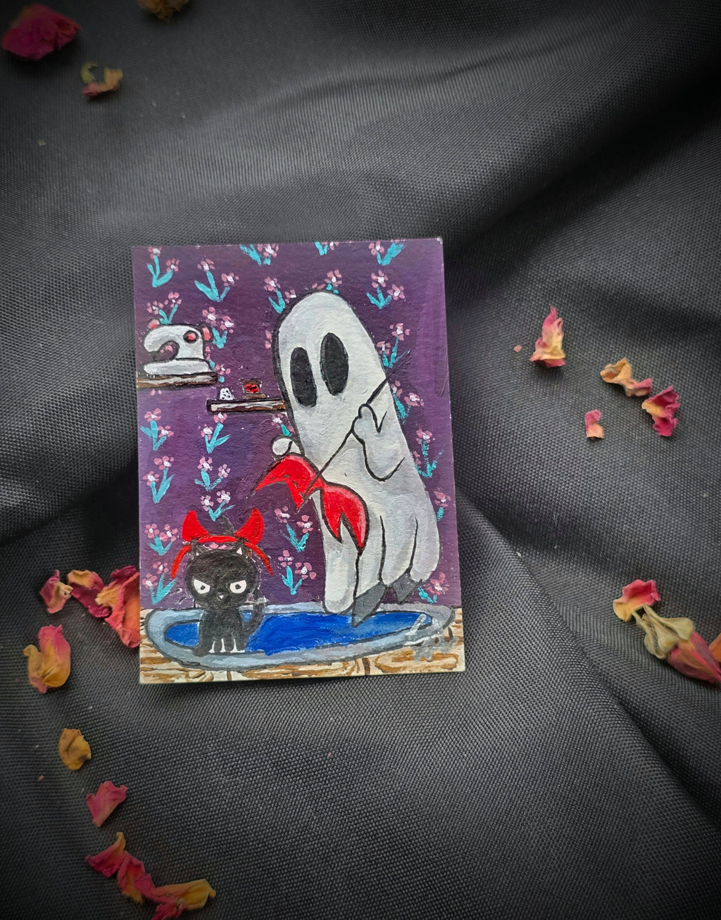 A Little Devil in the Making original ACEO painting featuring a white ghost sewing a red devil costume beside a black kitten wearing red devil horns, photographed on black fabric with dried rose petals.