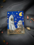 A Little Forever original Ghost and Kitty ACEO painting showing a white ghost holding flowers beside a black cat and gravestone, styled on black fabric with dried rose petals.