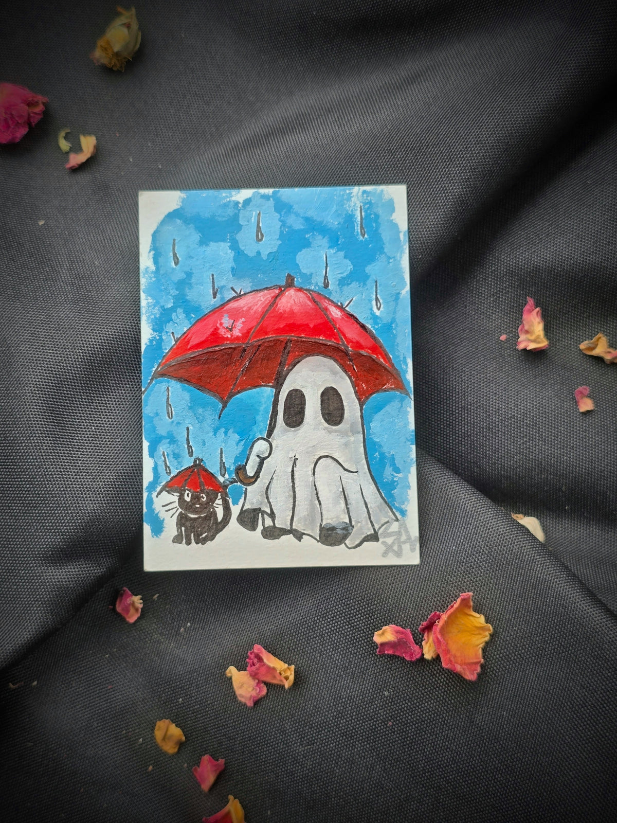 Original A Little Under the Weather ACEO painting featuring a white ghost holding a red umbrella beside a black cat with a tiny matching umbrella in the rain, photographed on black fabric with dried rose petals.