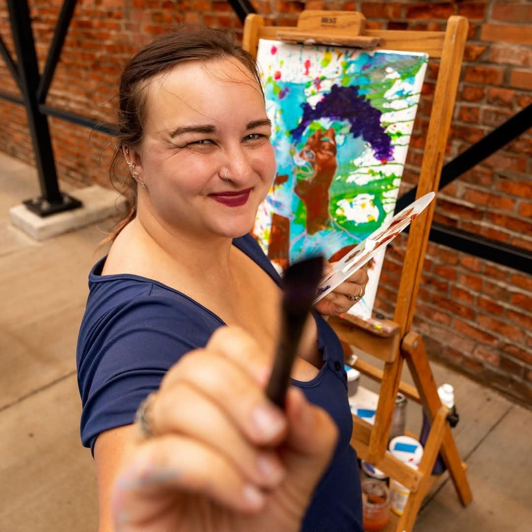 Artist holding a paintbrush beside a colorful live painting at an event