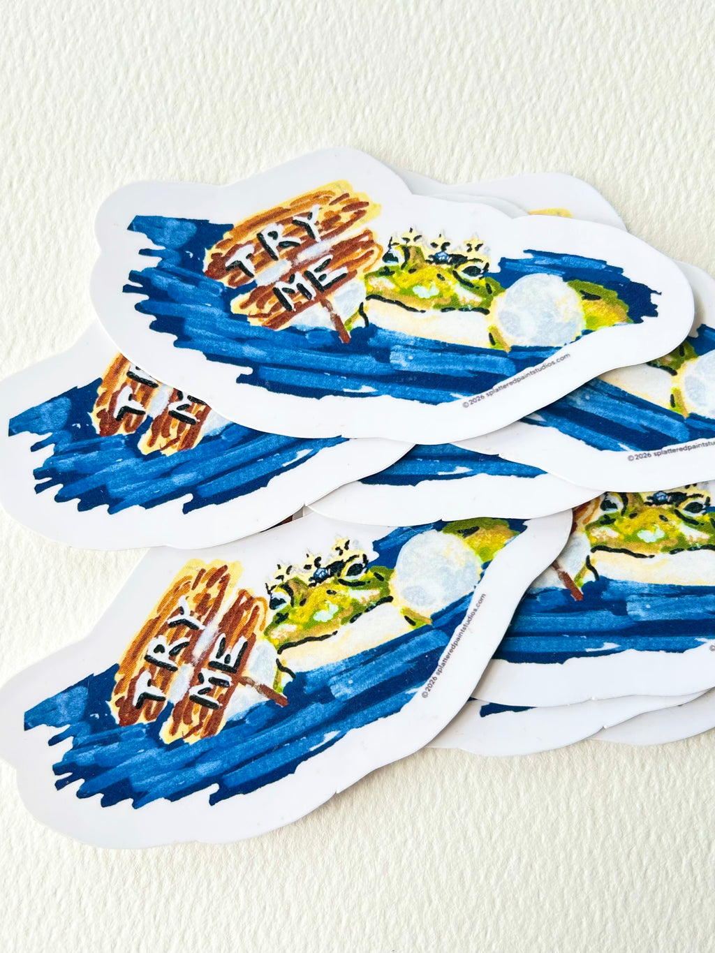 Stack of Try Me vinyl stickers featuring a crowned frog beside a painted Try Me sign