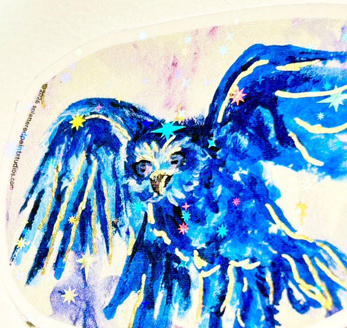 Detail of Voiding Radiance laminated vinyl sticker showing a blue bird with gold accents and holographic stars.