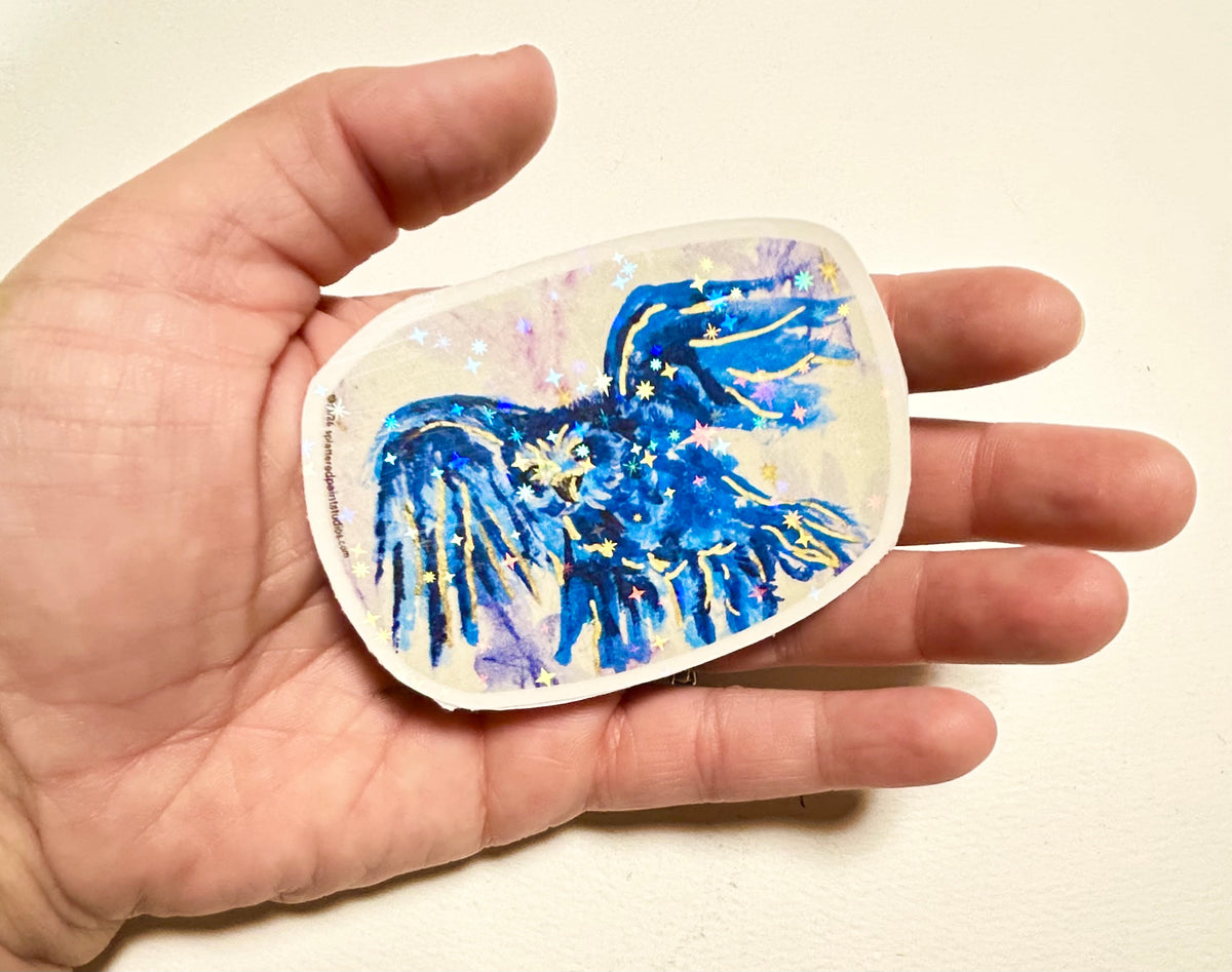 Voiding Radiance laminated vinyl sticker held in hand, showing the blue bird design with gold and holographic star details.
